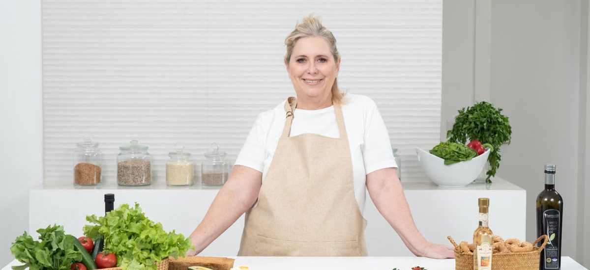 A mature woman in an apron stands at a kitchen counter with fresh produce and cheese, ready for cooking.
