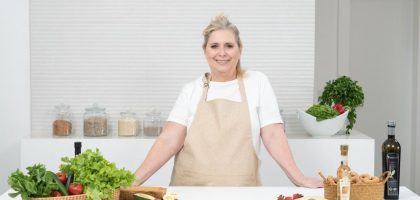 A mature woman in an apron stands at a kitchen counter with fresh produce and cheese, ready for cooking.