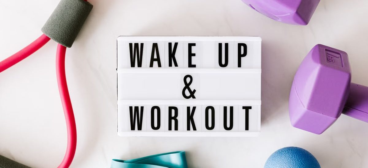 Top view closeup composition of wake up and workout inscription on light box surface among expanders and dumbbells and massage ball on white background